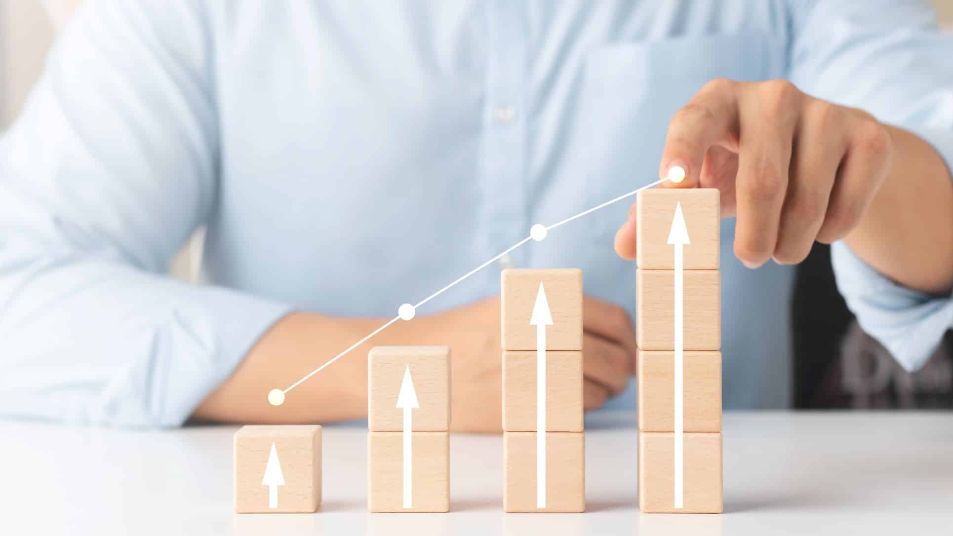 Investor businessman pointing to the top of increasing amount of blocks with up-arrows on it to show investment growth and success 