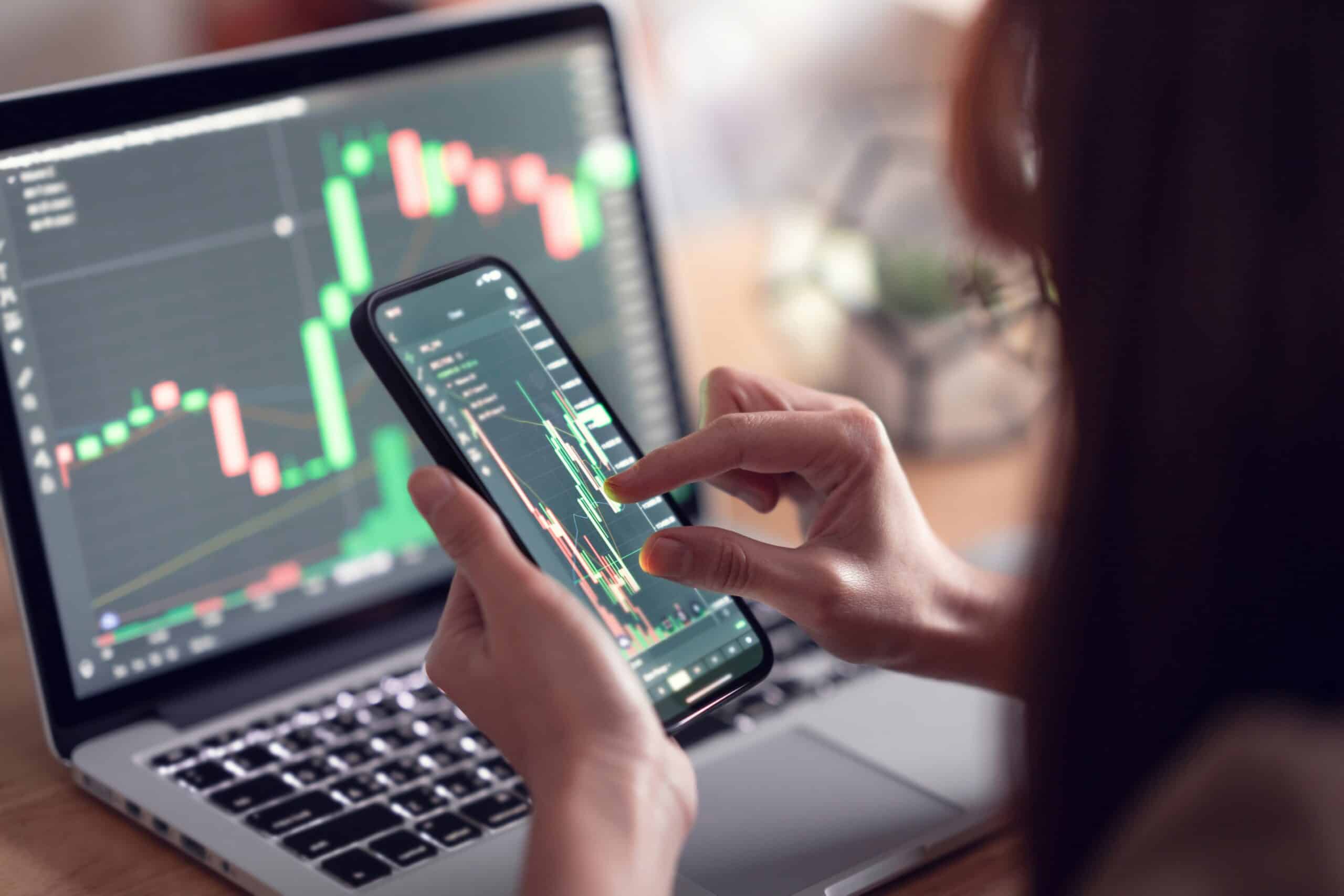 An investor looking at a fluctuating graph on her laptop and smartphone, showing the volatility of cryptocurrency.  