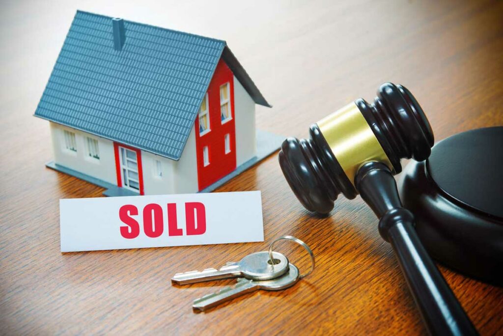 Small investment property house on a table next to a “sold” sign, keys, and judge’s gavel to signify the rules that govern Self-Directed IRA non-recourse loans.