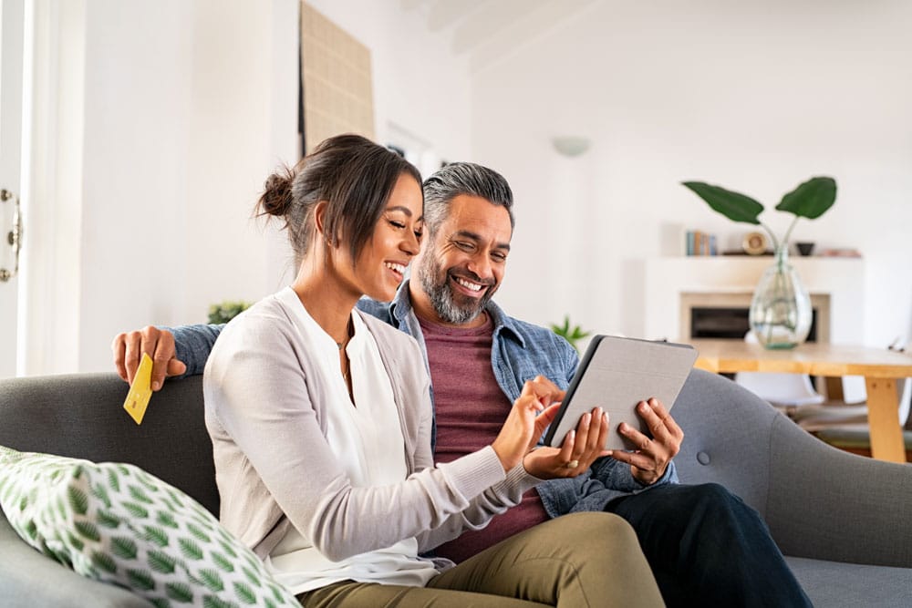 IRA LLC investor couple smiling on a couch looking at their tablet with their retirement savings on it.