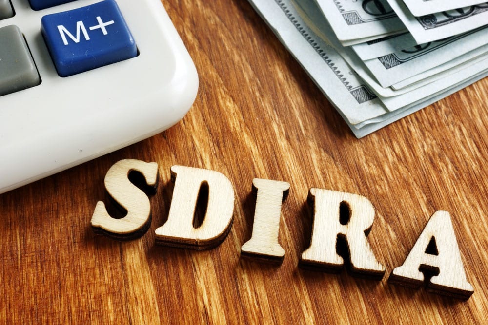 Letters on a desk that spell out “SDIRA” next to a calculator and money to show that you can grow your retirement savings with a Self-Directed IRA.