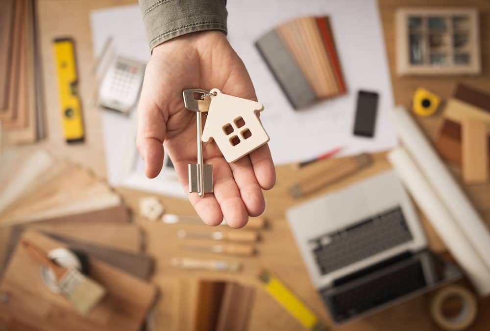 Hand holding a key with a miniature house keychain with a laptop, pencils, papers, and other tools on the table underneath to indicate an investment in commercial real estate with an IRA LLC.