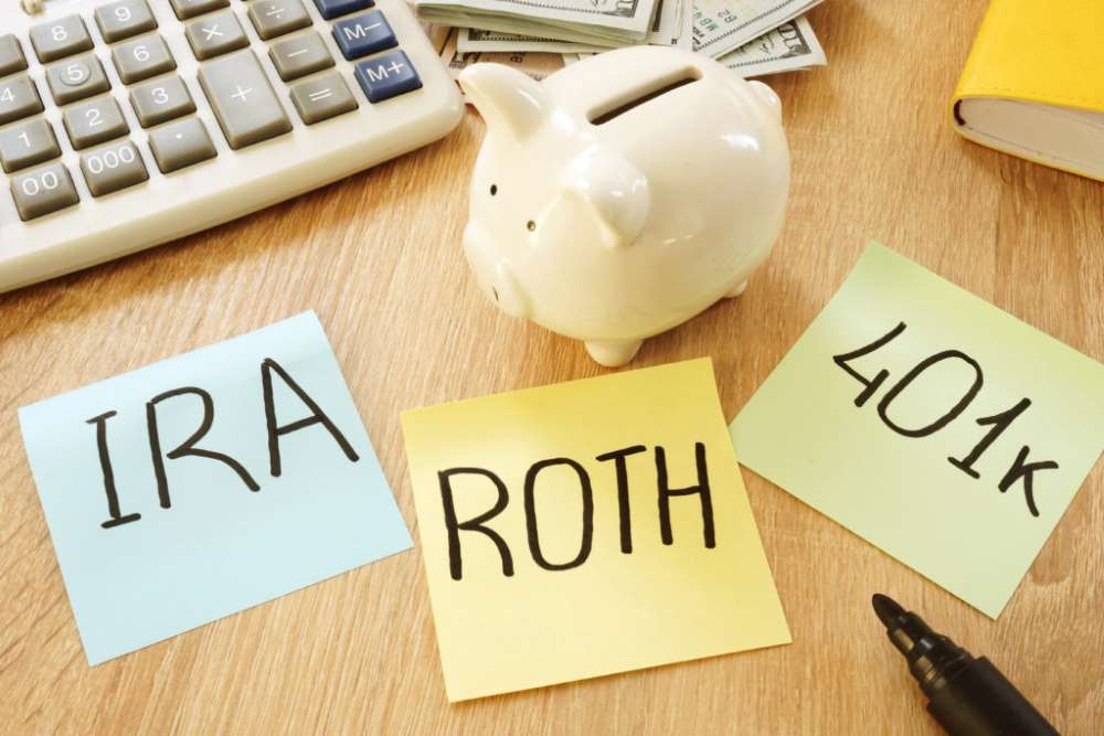 Three post-it notes with the words ‘Roth’, ‘401k’, and ‘IRA’ stuck on desk beside a piggybank, indicating that all these accounts are excellent options for retirement savings. 