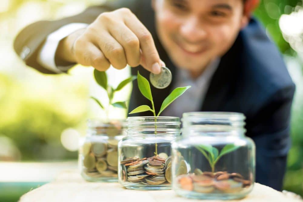 A man piles coins in his savings jars with plants growing outside of them, showcasing with patience and due diligence, growth in your retirement savings can occur.